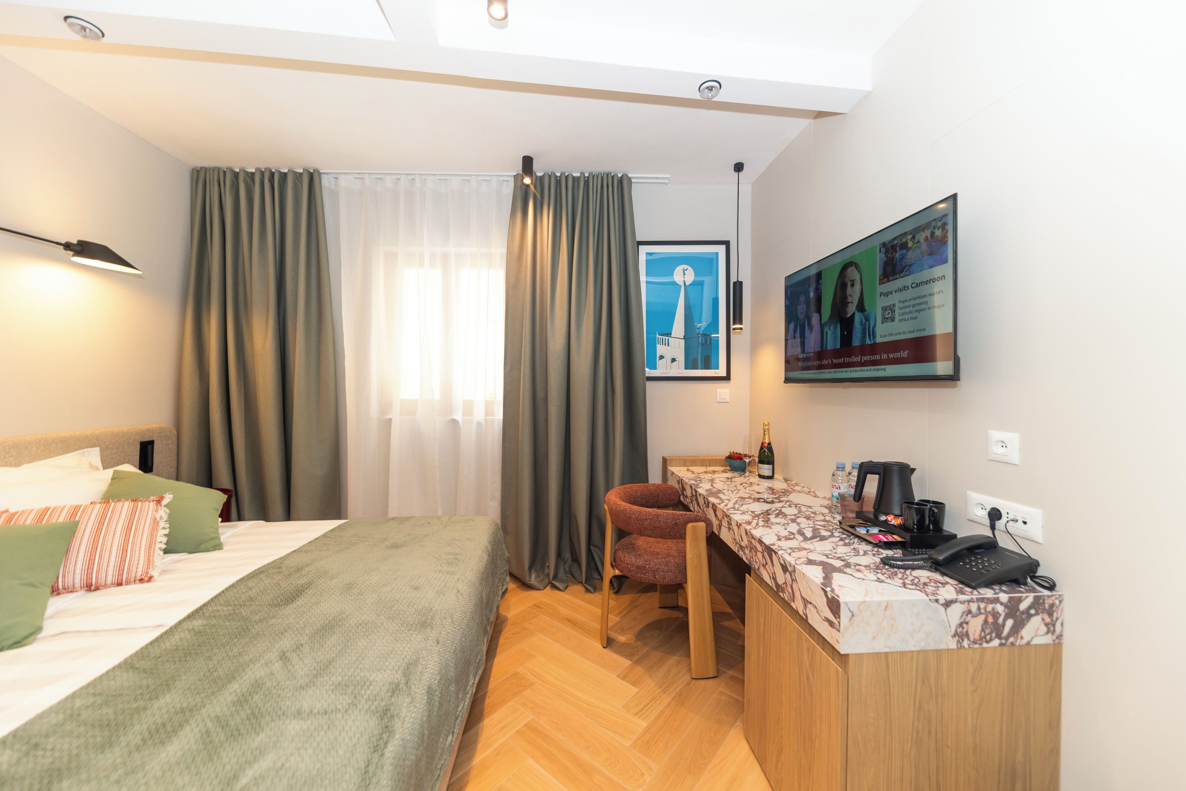 Habitación de hotel con cama, escritorio, silla, ventana con cortinas, un cuadro en la pared y un cartel.