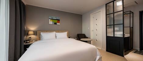 Quarto de hotel com uma cama grande, uma escrivaninha com luminária, uma cadeira, uma mesinha e um guarda-roupa com espelho.