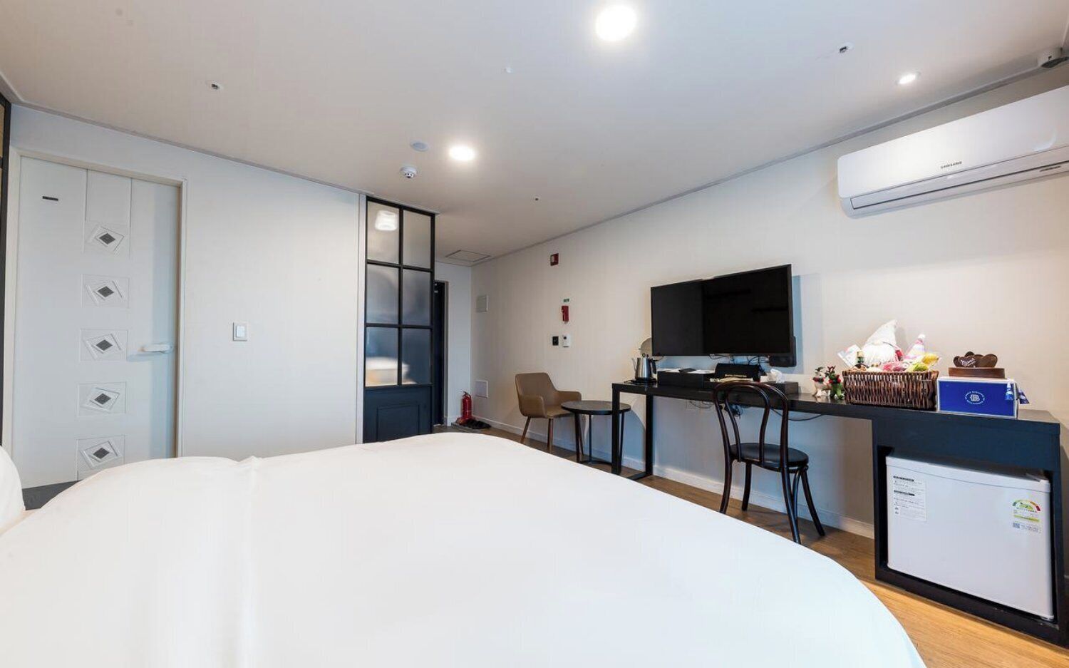 A modern hotel room with a large bed, a desk with a computer, a TV, and a small fridge.