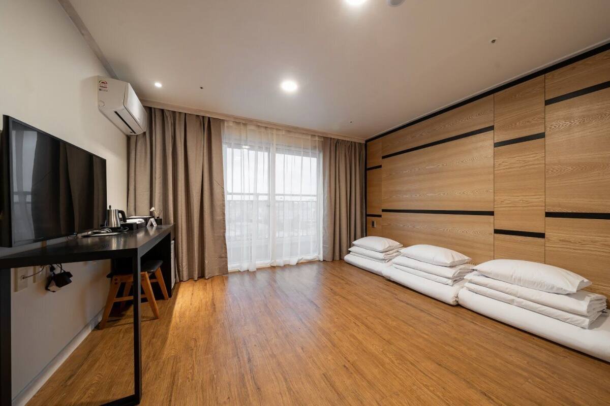 Una habitación moderna con piso de madera, televisor de pantalla plana, escritorio con silla, ventana grande con cortinas translúcidas y cama con almohadas.