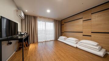 A modern room with a wooden floor, a flat-screen TV, a desk with a chair, a large window with sheer curtains, and a bed with pillows.