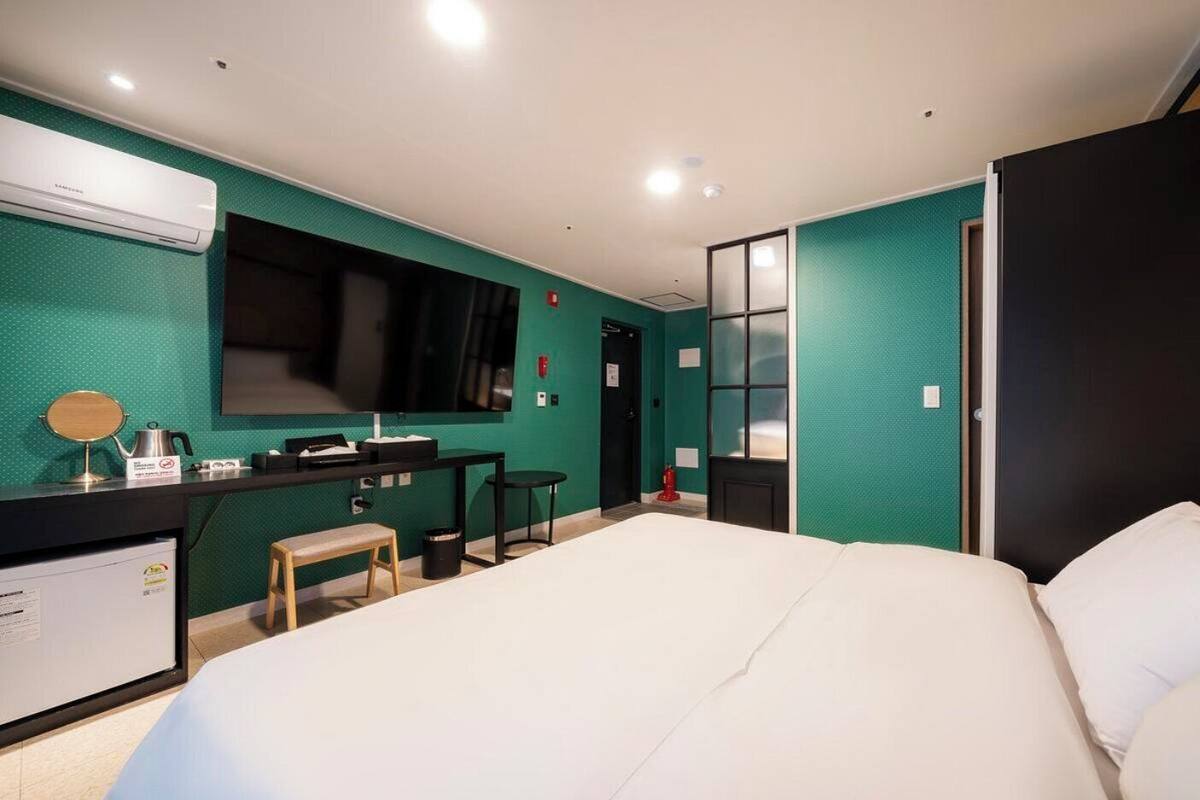 Habitación de hotel con cama, escritorio con espejo, taburete pequeño, cesto de basura y aire acondicionado de pared.