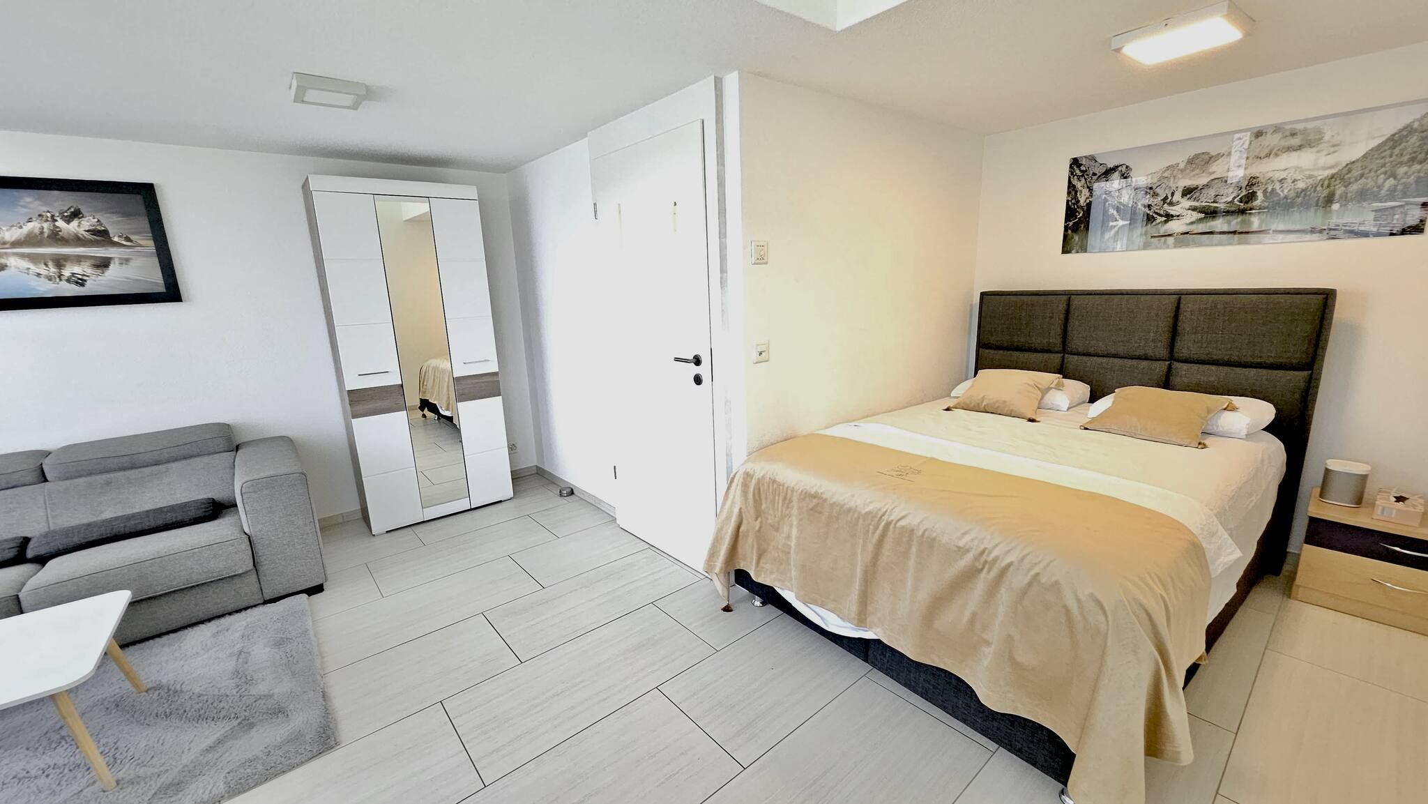 A bedroom with a bed, a grey sofa, a small table, a wardrobe, and a painting on the wall.