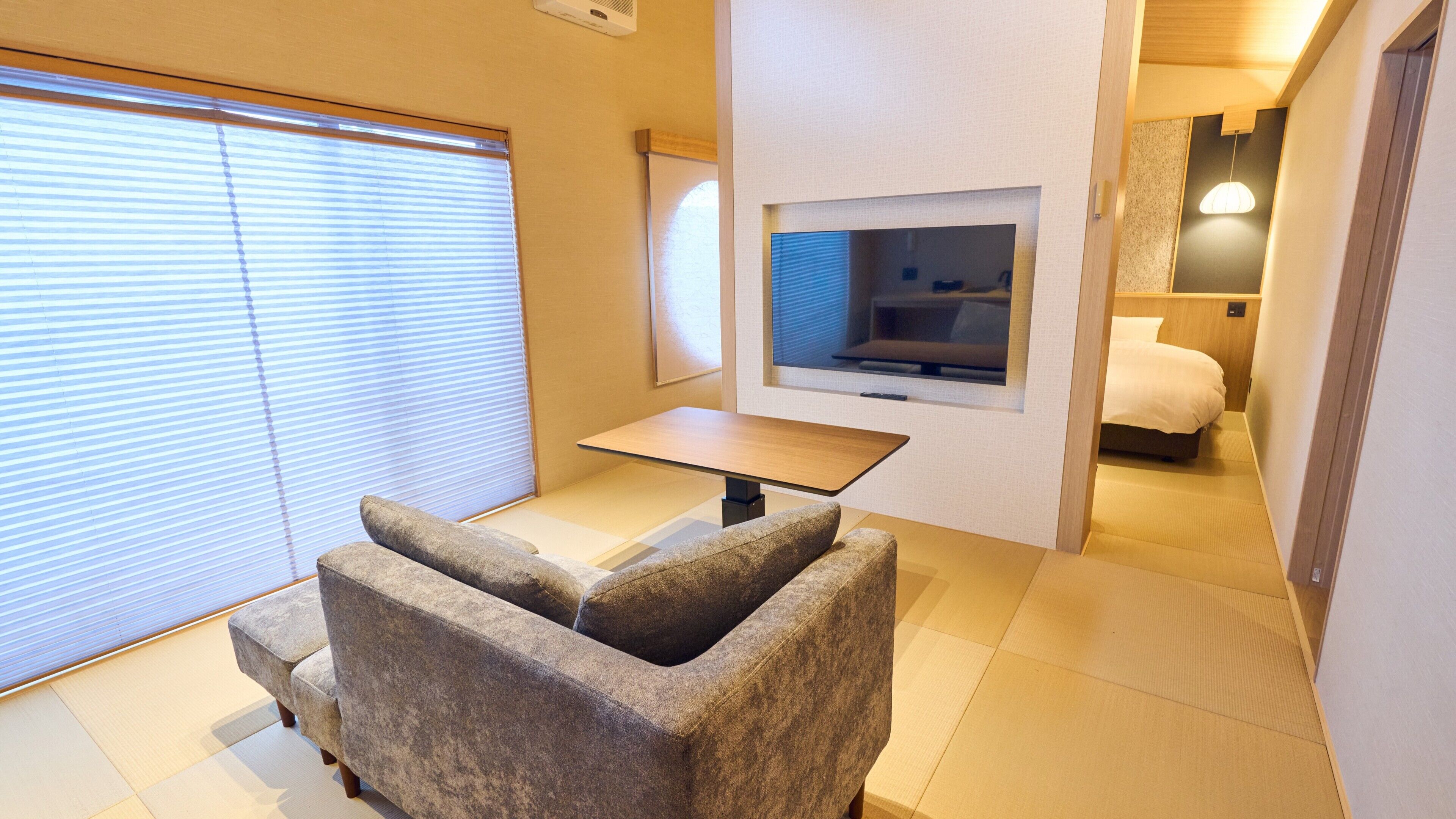A hotel room with a sofa, a small table, a TV mounted on the wall, and a bed with a wooden headboard.