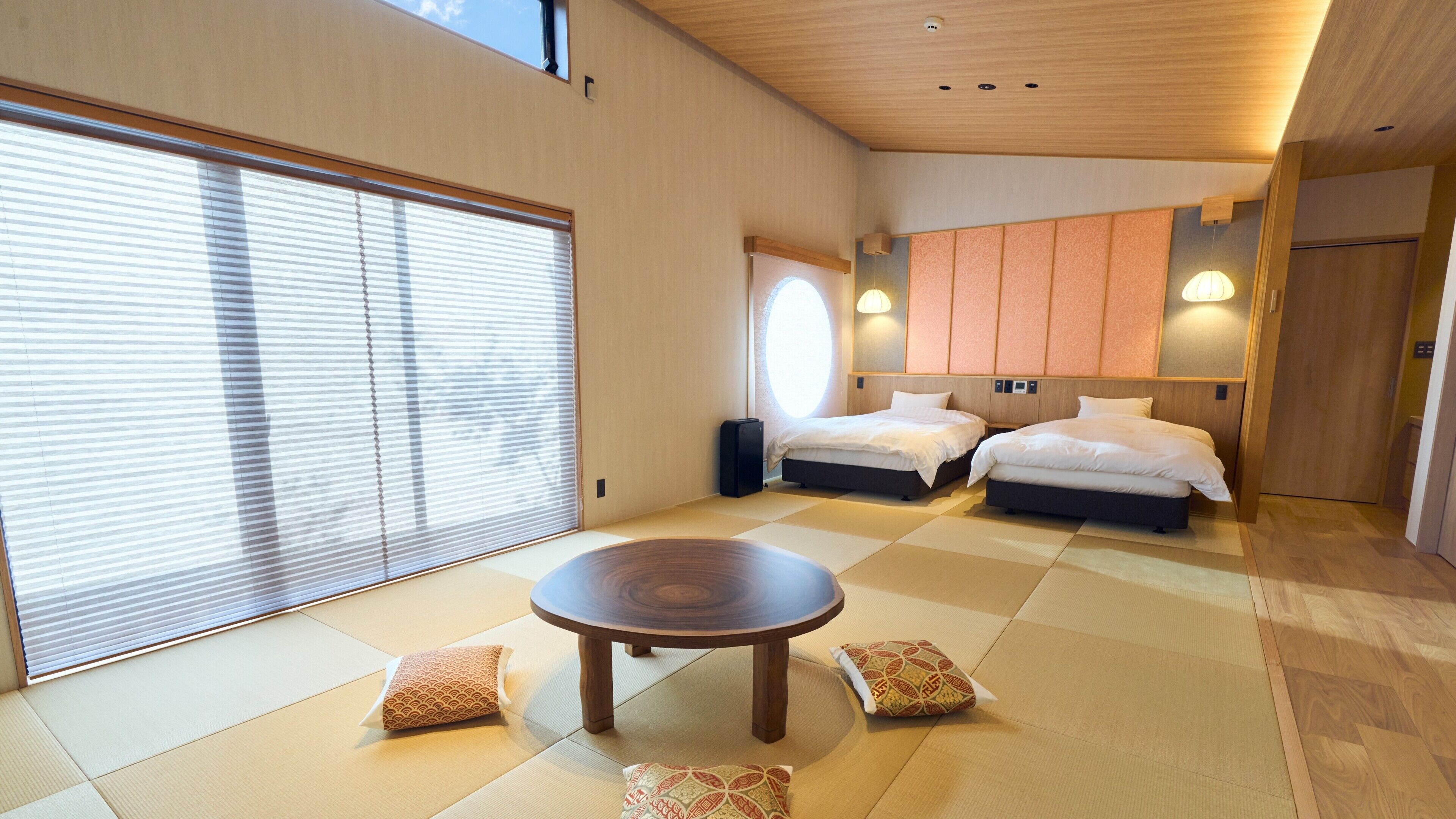 A modern hotel room with tatami flooring, two beds, a round wooden table, and a large window with blinds.