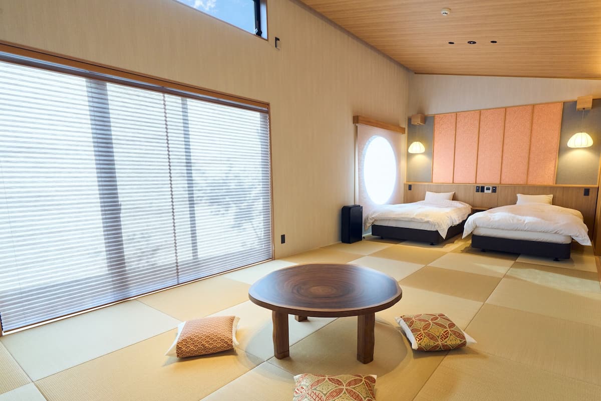 Una habitación de hotel moderna con suelo de tatami, dos camas, una mesa redonda de madera y un ventanal con persianas.