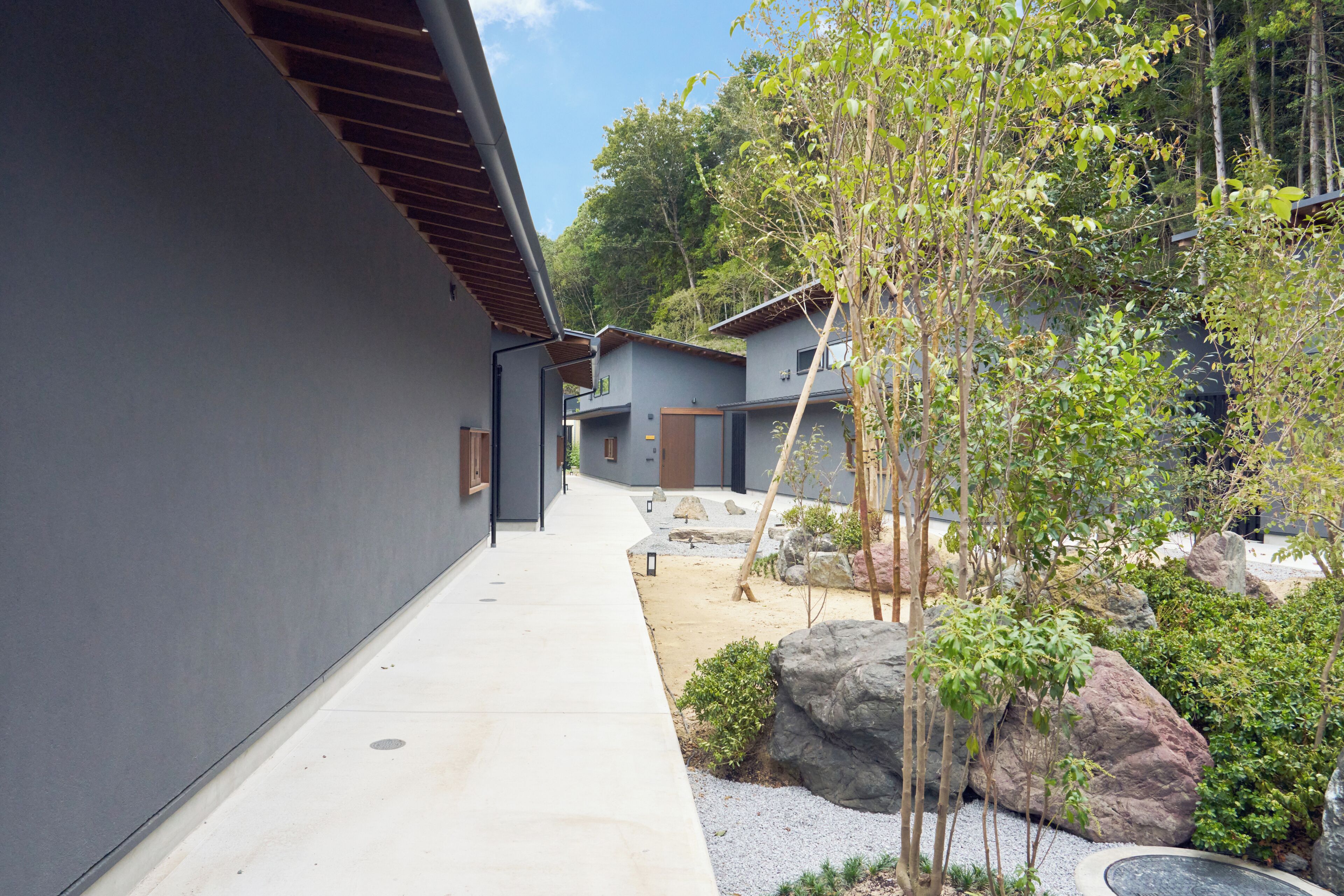 A modern building with a concrete exterior, a paved walkway, and a landscaped garden with rocks and trees.