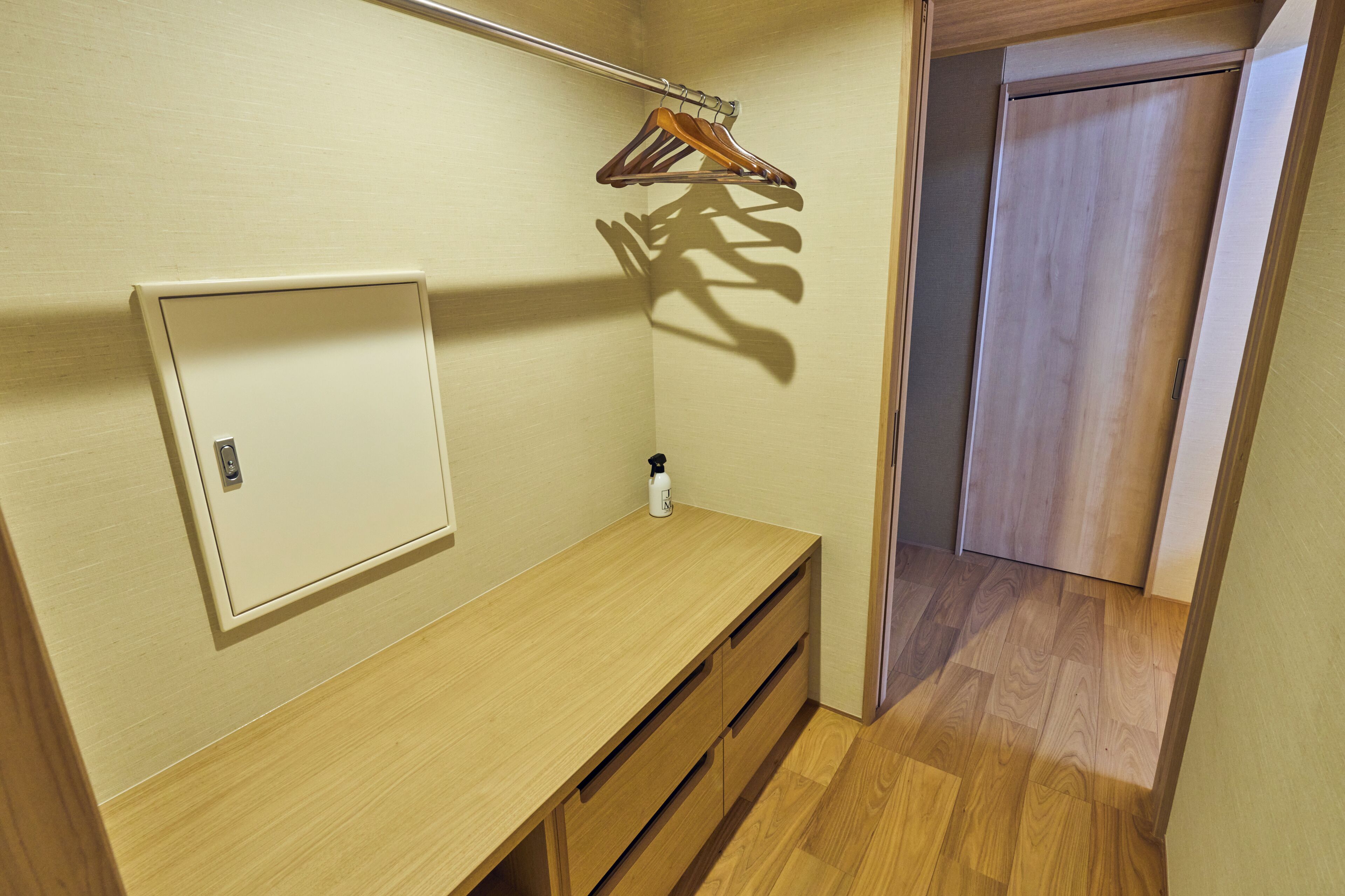 A room with a wooden desk, a hanging clothes rack, a small shelf with a bottle, and a door leading to another room.