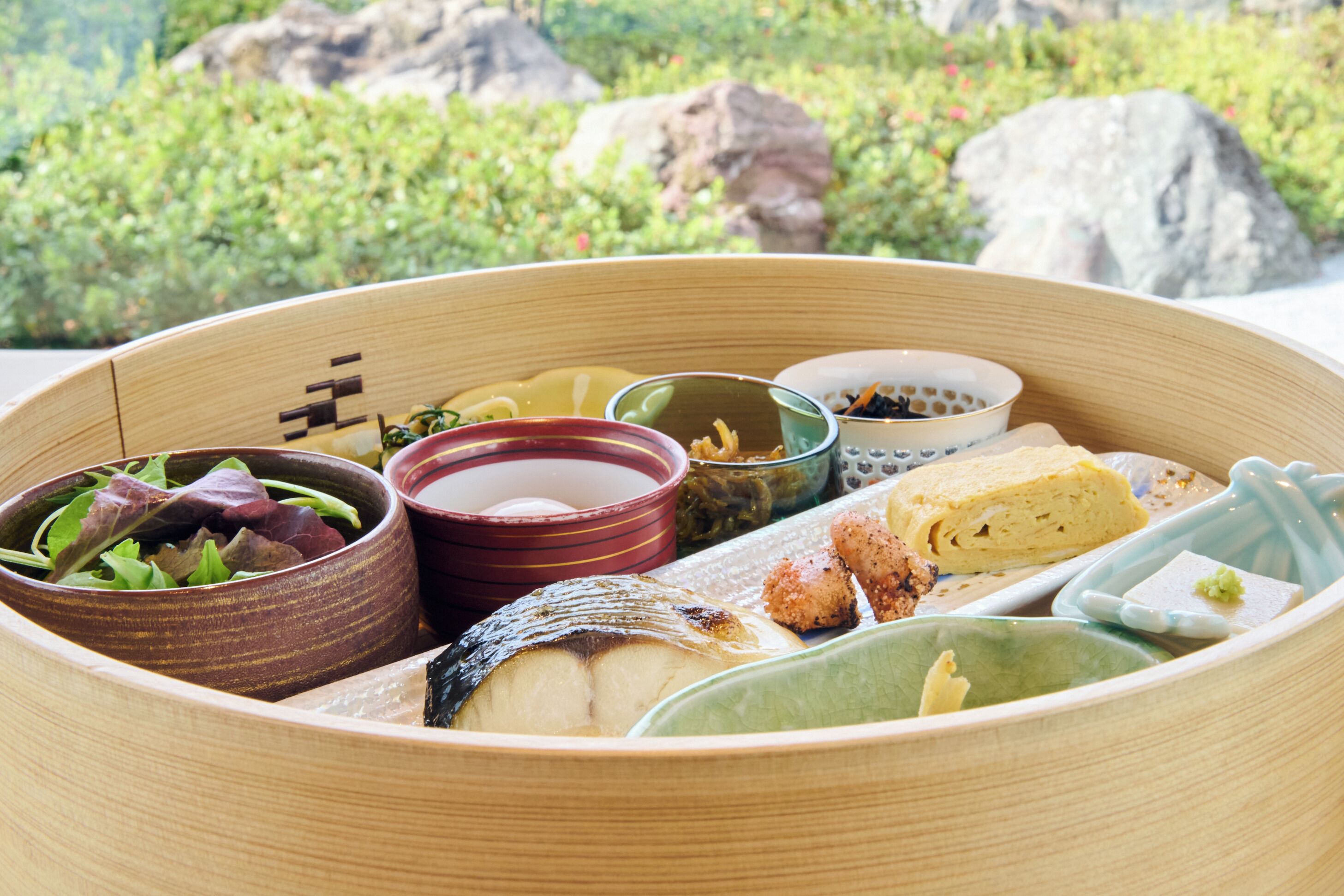 Eine traditionelle japanische Bento-Box mit verschiedenen Speisen und kleinen Schalen, vor einer natürlichen Kulisse.