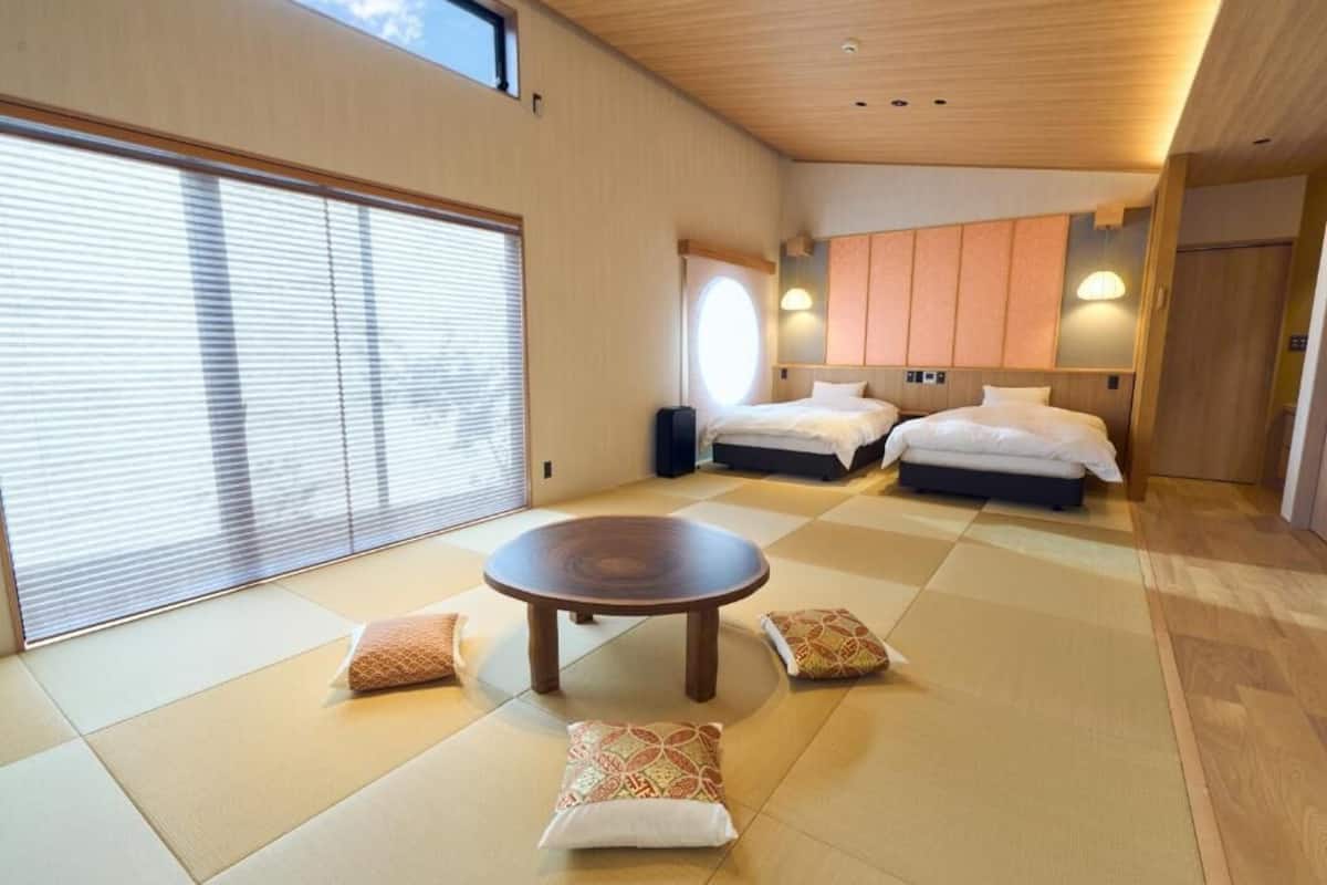 Una habitación con suelo de tatami, dos camas, una mesa redonda y un amplio ventanal con persianas.