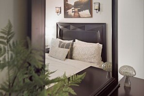 Ein Schlafzimmer mit dunklem Kopfteil, einem Bett mit gemusterten Betttextilien, zwei Wandlampen und einem gerahmten Bild über dem Bett.
