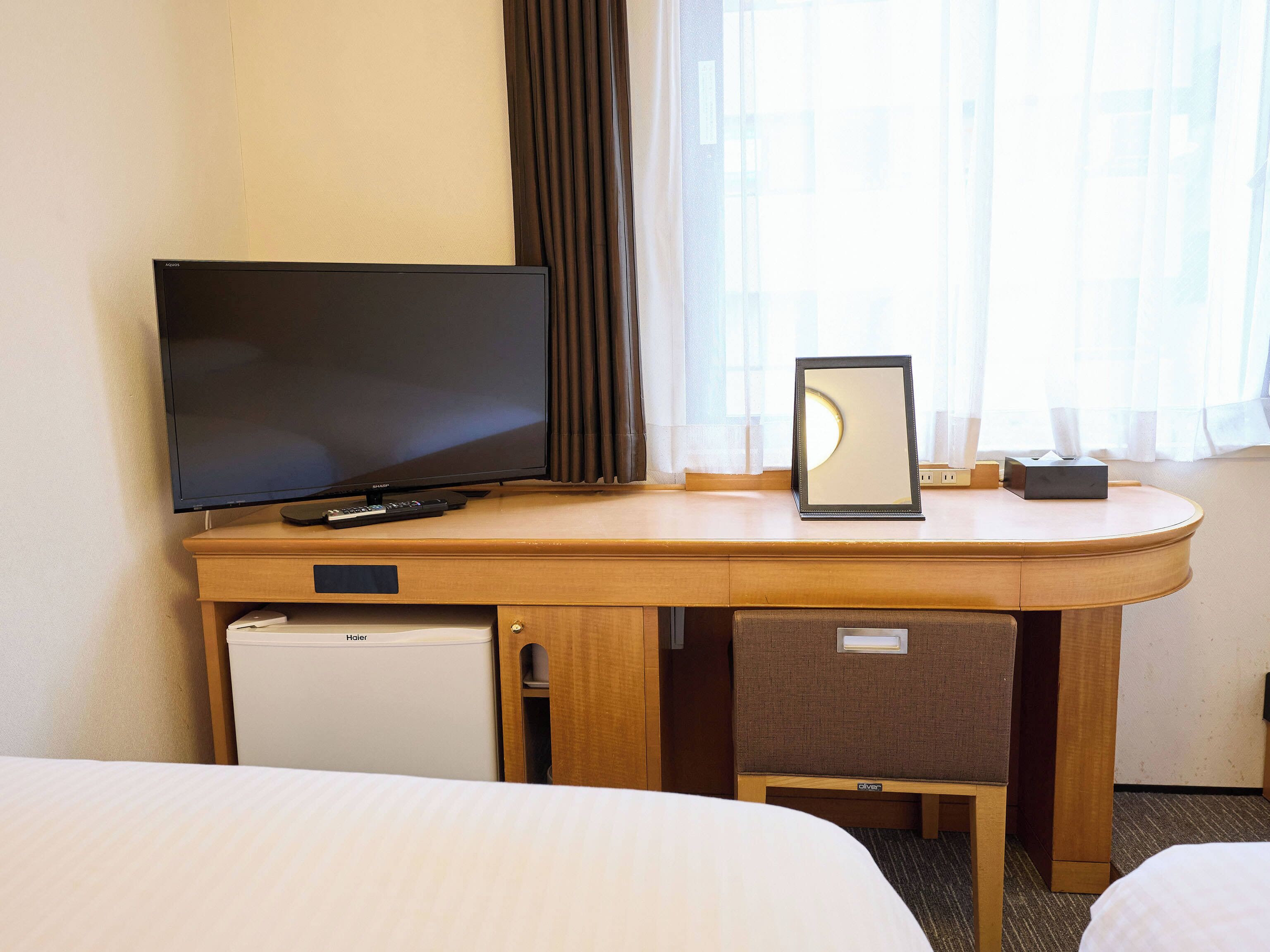 A hotel room with a desk, television, mirror, and a small refrigerator.