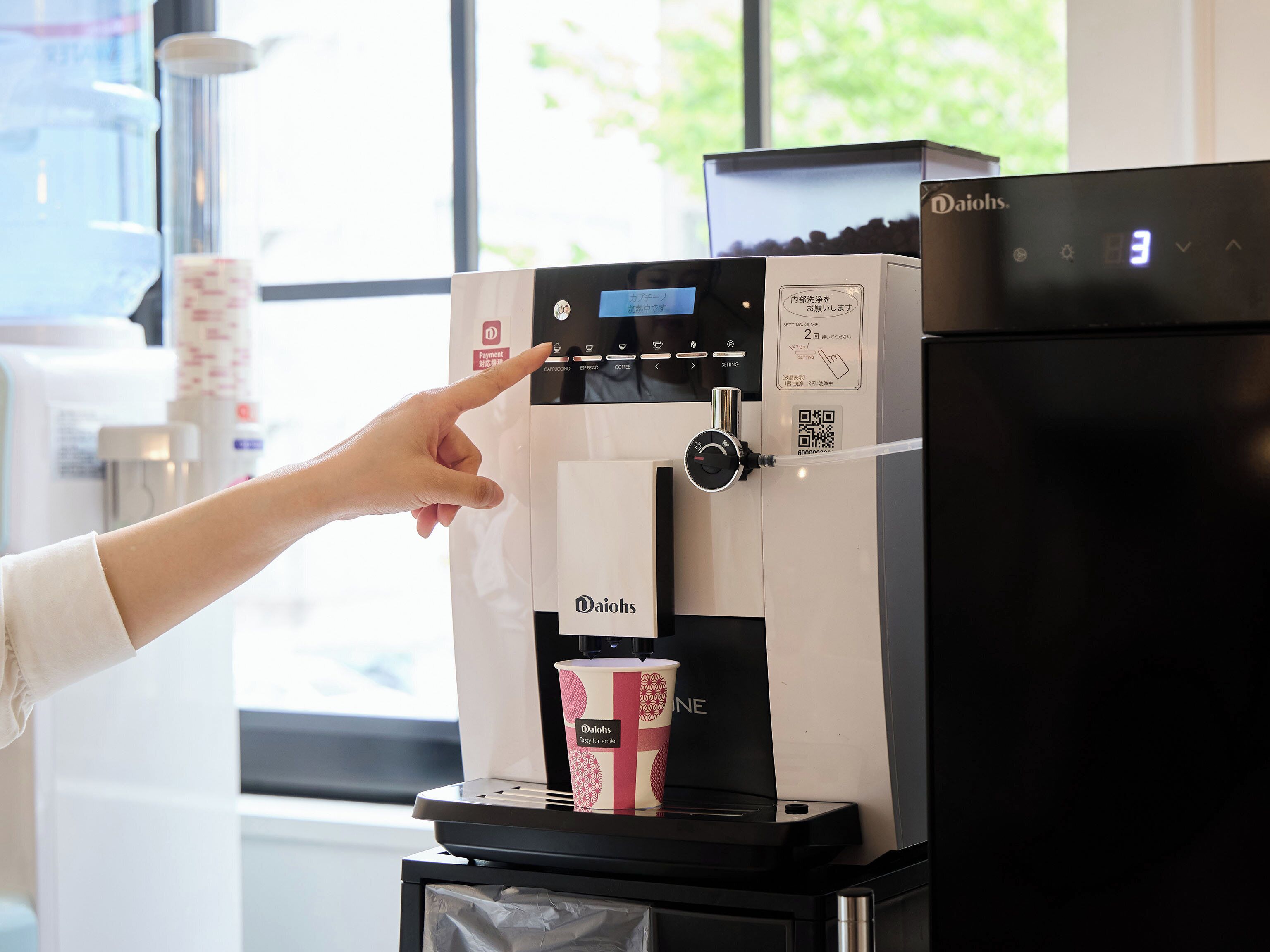 A coffee machine with a digital display, a payment button, and a cup holder for a branded cup.