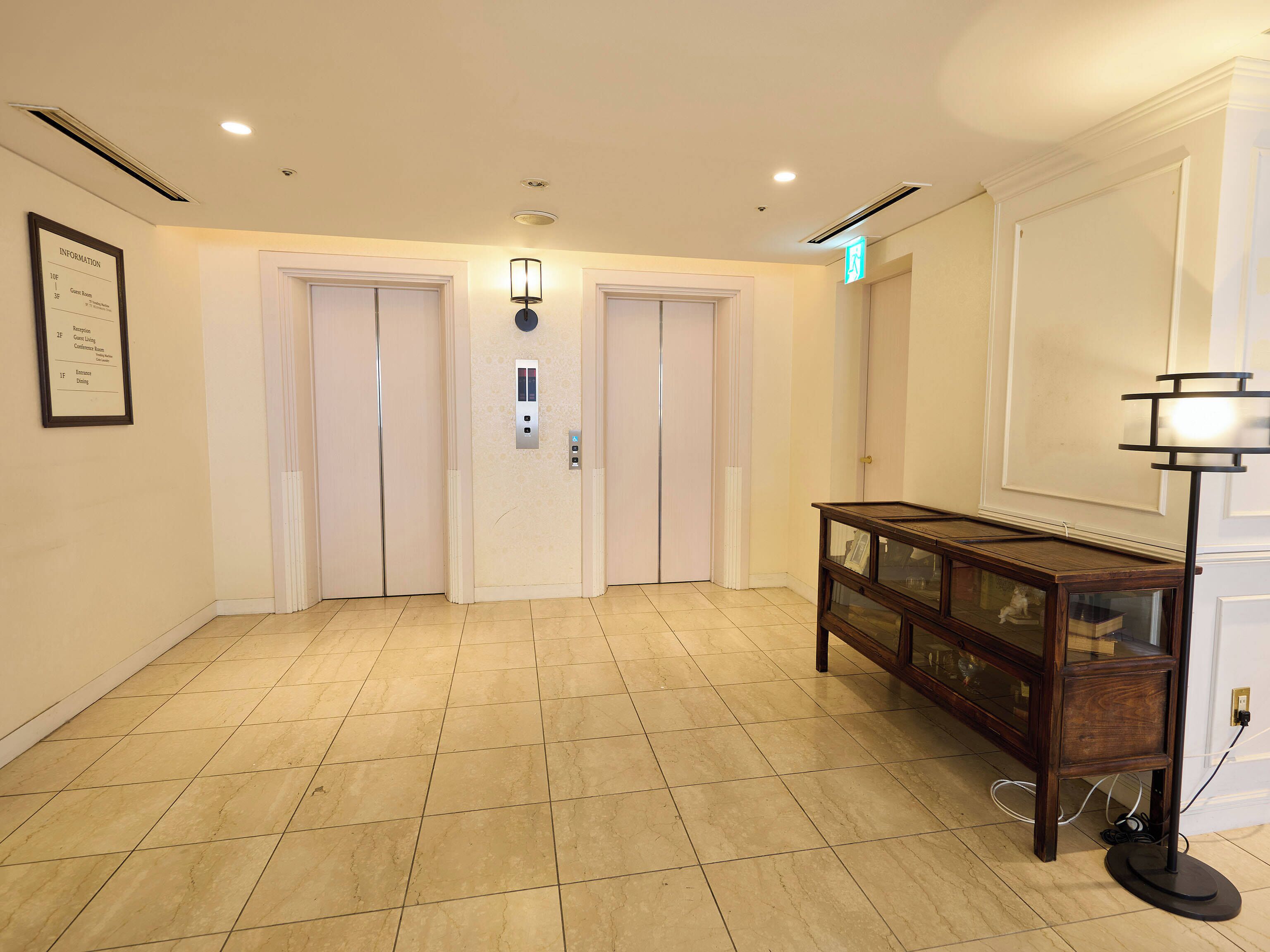 A hotel lobby with a reception desk, two elevators, and a wall-mounted information sign.
