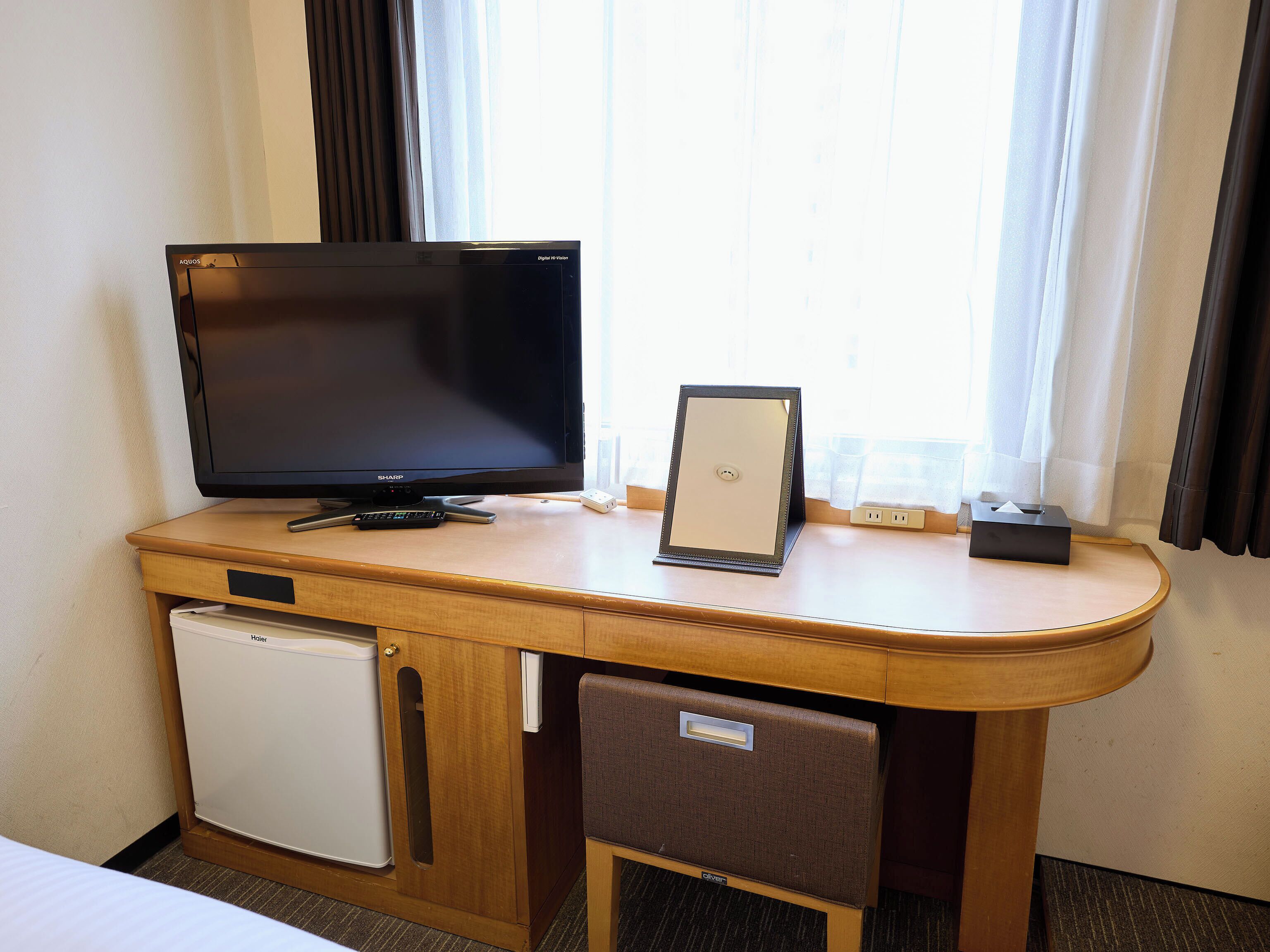 A hotel room with a wooden desk, a television, a mini-fridge, a chair, and a window with curtains.