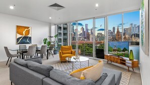 Un salón moderno con vistas a la ciudad, sofás grises, un sillón amarillo y un comedor con una mesa y sillas de vidrio.