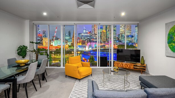 Una sala de estar moderna con vistas a la ciudad, un comedor con mesa de vidrio y un televisor sobre un mueble de madera.
