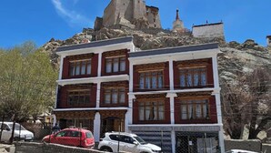 Um edifício com arquitetura tradicional, uma estrutura imponente semelhante a uma fortaleza ao fundo, e um céu azul claro.