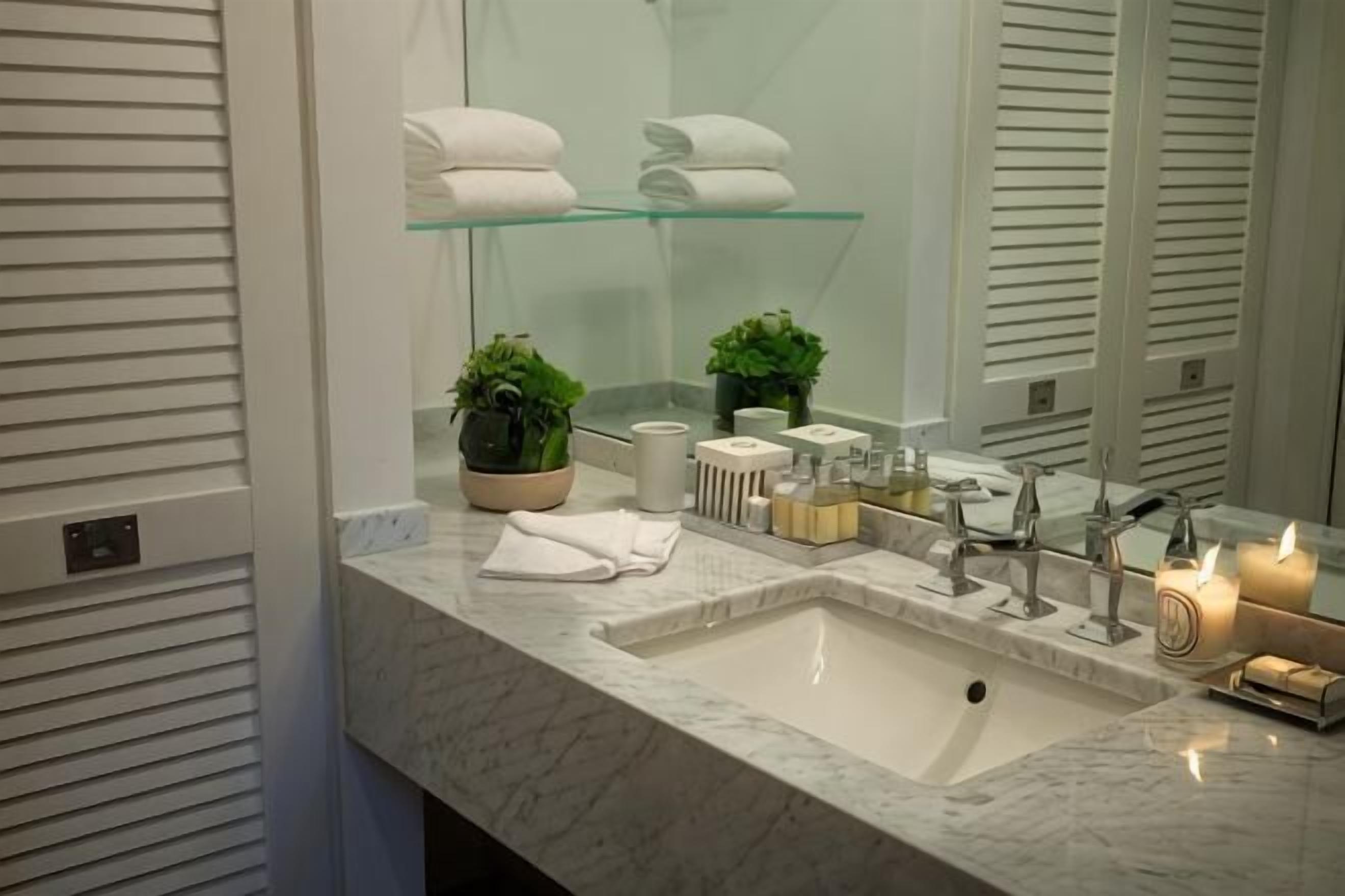 Un baño con lavamanos de mármol, estantes de vidrio, una planta en maceta y artículos de aseo.