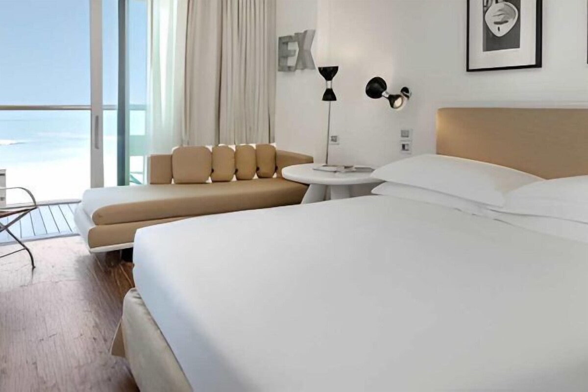 Habitación de hotel con una cama grande, un sofá, una mesita redonda y vista al océano.