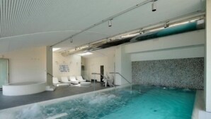 Una piscina interior con vista al agua cristalina, una zona de estar con sillas blancas y un área de spa con una bañera de hidromasaje circular.