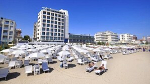 Una playa con filas de tumbonas y sombrillas blancas, un edificio de hotel alto y otros edificios al fondo.