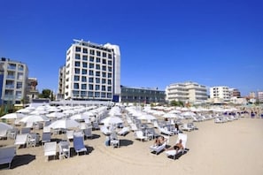 Una playa con filas de tumbonas y sombrillas blancas, un edificio de hotel alto y otros edificios al fondo.