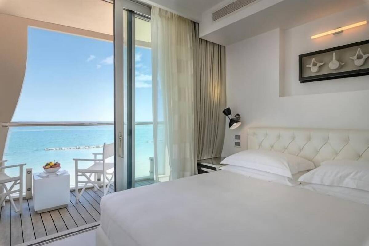 Habitación de hotel con cama, una mesita con fruta, una silla y balcón con vista al mar.