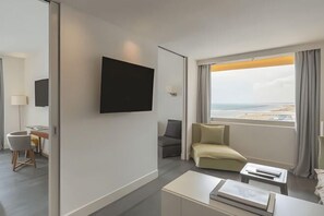 A hotel room with a flat-screen TV mounted on the wall, a sofa, a small table, and a window with a view of the beach. - Excelsior Hotel, Spa & Lido (Pesaro)