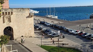 Una zona costera con un muro de piedra histórico, un puerto deportivo con barcos y un estacionamiento.
