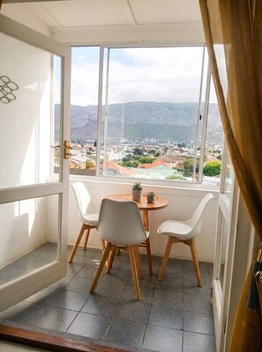 A balcony with a table and chairs, a view of a town and mountains, and a window with curtains. - Outspan Lodge- Giraffe House (Cape Town)