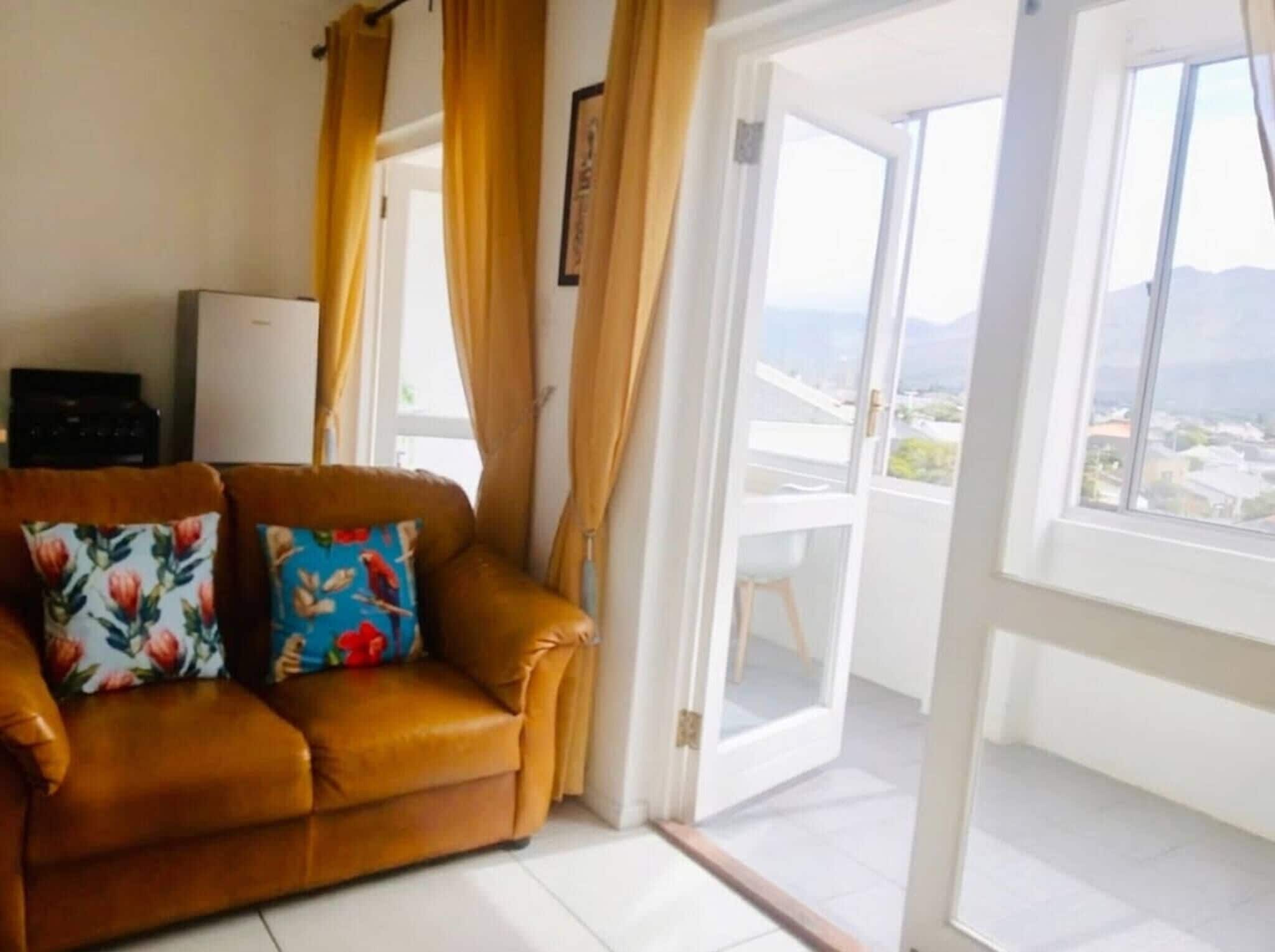 A living room with a brown leather sofa, two floral cushions, a small black appliance, and a sliding glass door leading to a balcony with a city view.