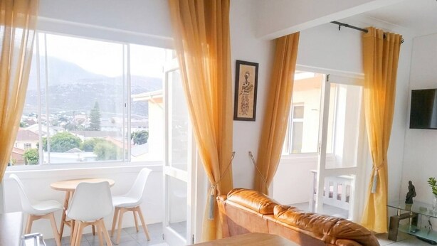 A modern living room with a sofa, a dining table, and chairs. There are large windows with curtains, a framed picture on the wall, and a TV mounted on the wall. - Outspan Lodge- Giraffe House (Cape Town)