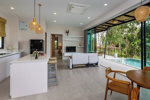 A modern kitchen with a central island, a dining area with a table and chairs, a TV, and a large window overlooking a pool. - HinKong Views - Tropical modern 2br sea view pool Villa - B (Tambon Ko Pha-ngan)