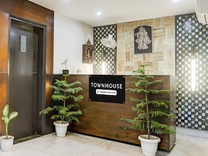 A reception desk with a sign reading 'TOWNHOUSE Company Serviced,' potted plants, a wall-mounted fan, and a decorative wall piece. - Townhouse Sushant Lok Phase 1 Hotel Neven (Gurugram)