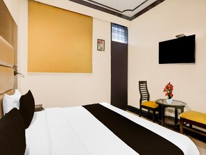 A hotel room with a bed, a chair, a small table with a vase of flowers, a window with bars, and a flat-screen TV mounted on the wall. - Townhouse Sushant Lok Phase 1 Hotel Neven (Gurugram)