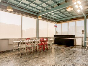 A room with a piano, tables, chairs, and large windows. - Townhouse Sushant Lok Phase 1 Hotel Neven (Gurugram)