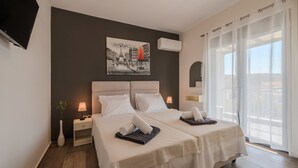 Ein Hotelzimmer mit zwei Betten, einem Bild an der Wand, einem Fernseher und einem Balkon mit Vorhängen.