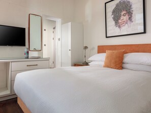 Ein Hotelzimmer mit Bett, Schreibtisch mit Flachbildfernseher, Spiegel, gerahmtem Kunstwerk und Nachttischlampe.