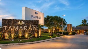 Entrada de hotel com uma grande placa, parede de pedra com logotipo de árvore e um caminho bem iluminado.