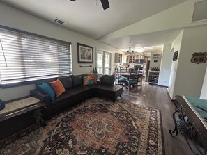 A living room with a sectional sofa, a patterned rug, a dining area with chairs, and a kitchen in the background.