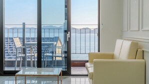 A living room with a sofa, a glass table, and two chairs, overlooking a balcony with a view of the sea.
