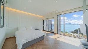 A bedroom with a large bed, a small table, and a view of the sea through a sliding glass door.