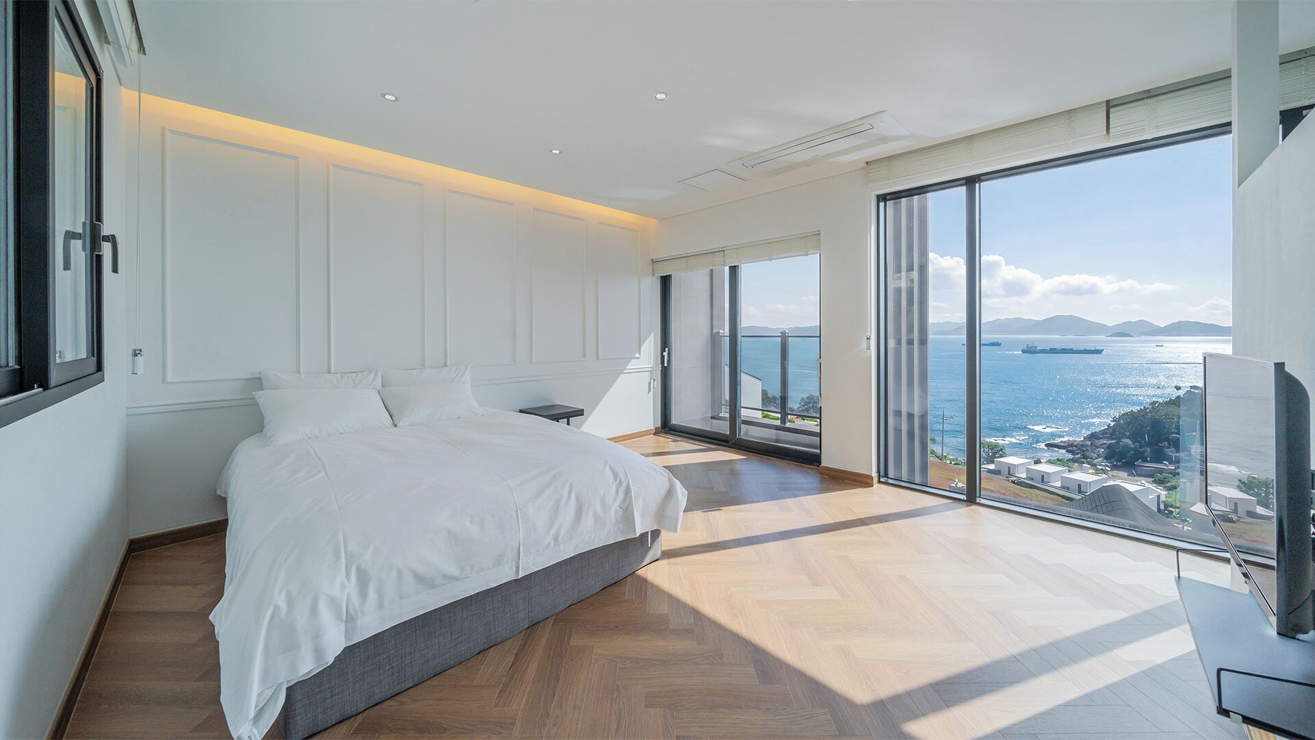 A modern bedroom with a large bed, a small table, and a TV, featuring a large sliding glass door leading to a balcony with a sea view.