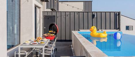 A rooftop pool with a yellow inflatable duck, a barbecue grill, and a dining table with food.