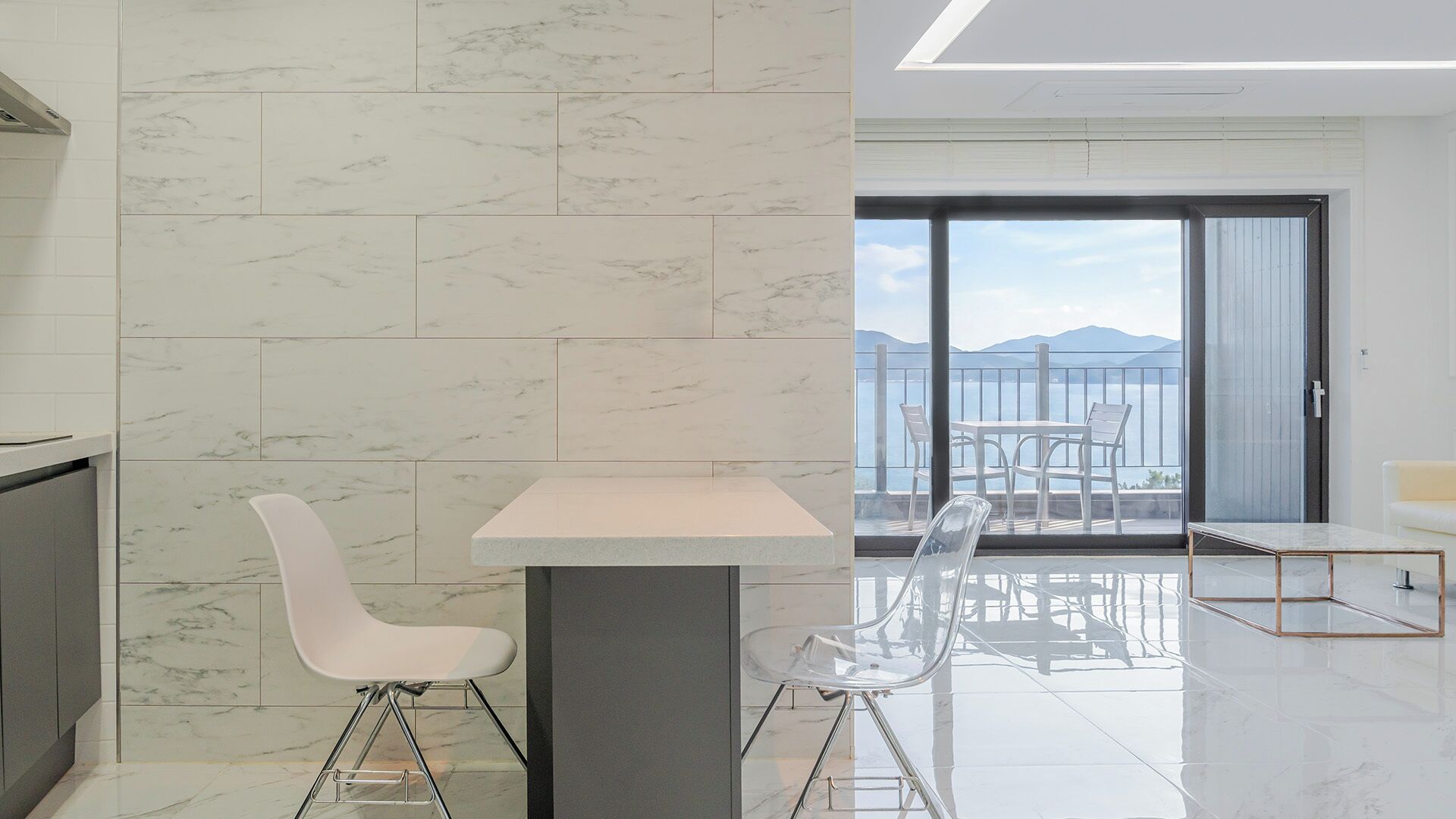 A modern kitchen with white marble tiles, a white dining table with two chairs, and a view of the sea through a sliding glass door.