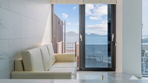A modern living room with a sofa, a glass table, and a view of the sea through a large sliding door.