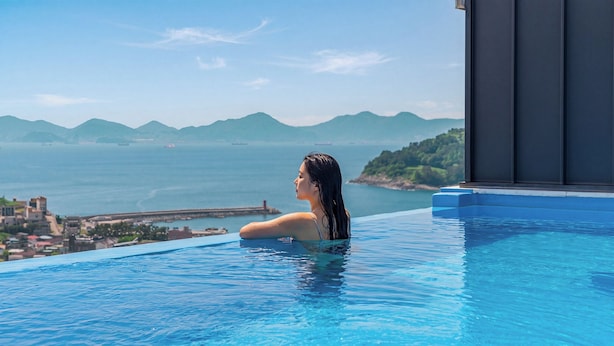 A person in a clear blue infinity pool with a view of the sea and distant mountains.