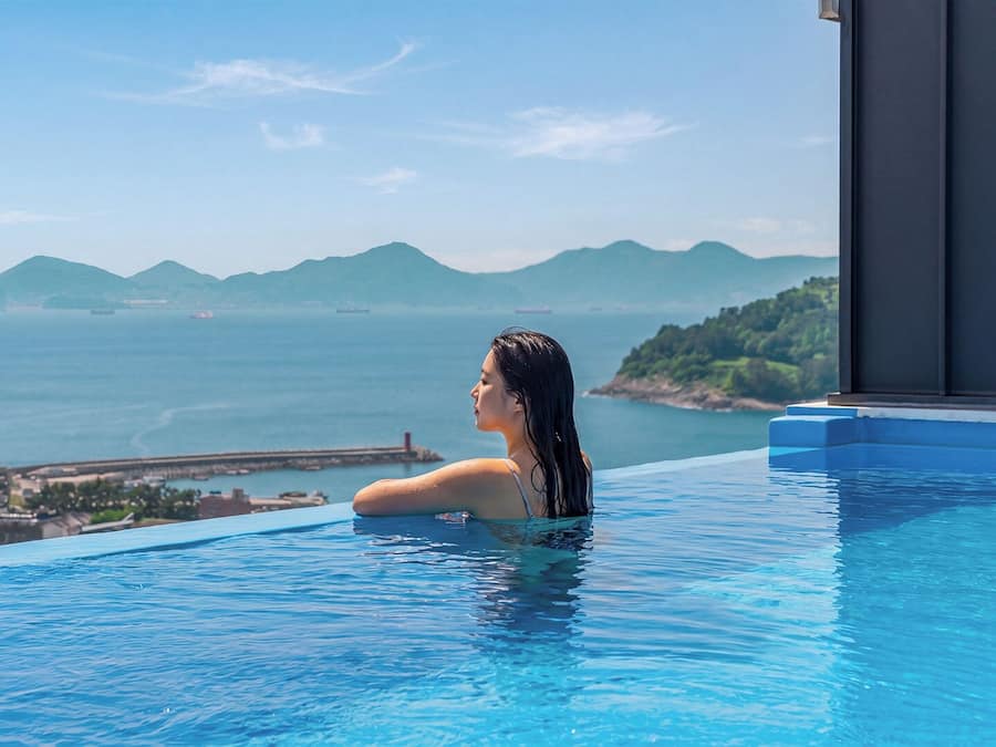 Eine Person in einem türkisfarbenen Infinity-Pool mit Blick auf das Meer und ferne Berge.