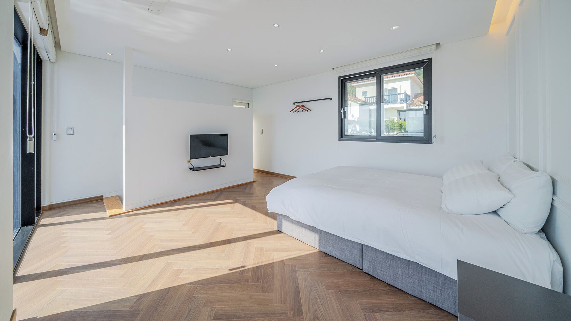 Une chambre moderne avec un grand lit, un téléviseur fixé au mur et une fenêtre donnant sur des immeubles.